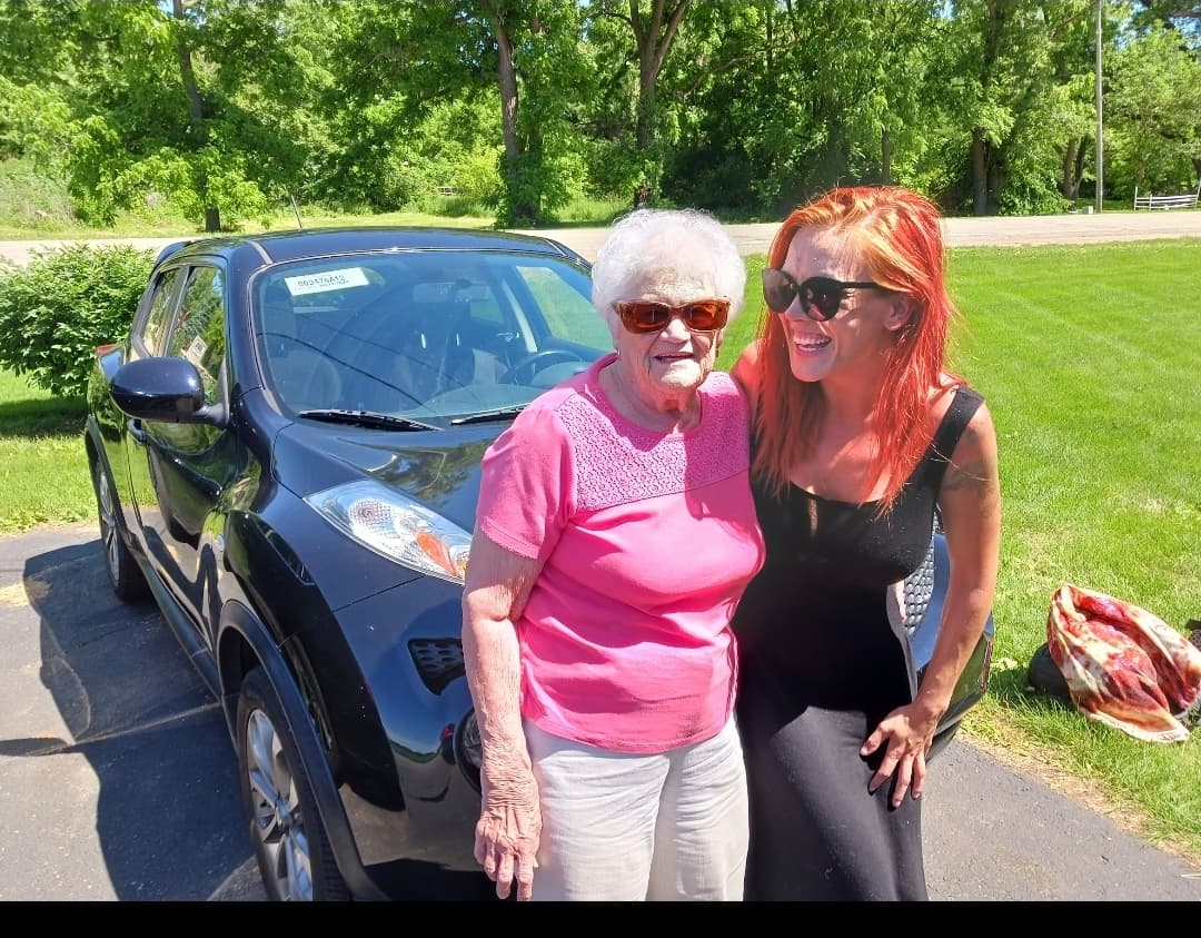 Smiling elderly woman in pink and laughing red-haired woman standing before a black SUV.