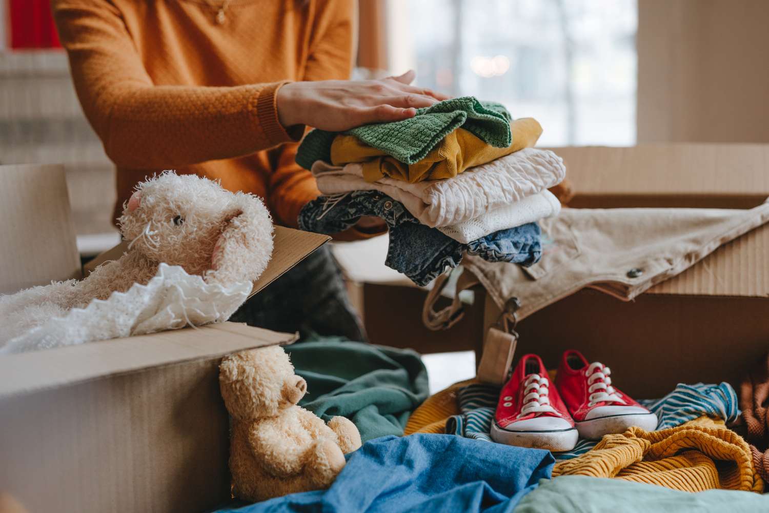 Person in orange sweater packing clothes, stuffed animals, and red sneakers into cardboard boxes.
