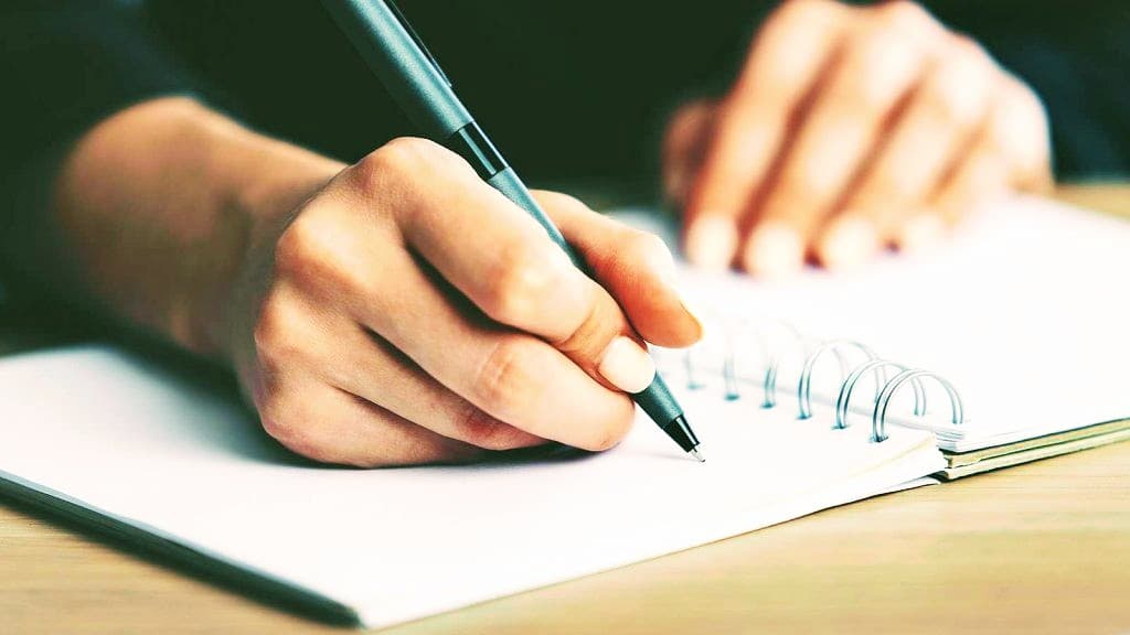 Close-up of a hand holding a black pen, writing in a white spiral notebook.