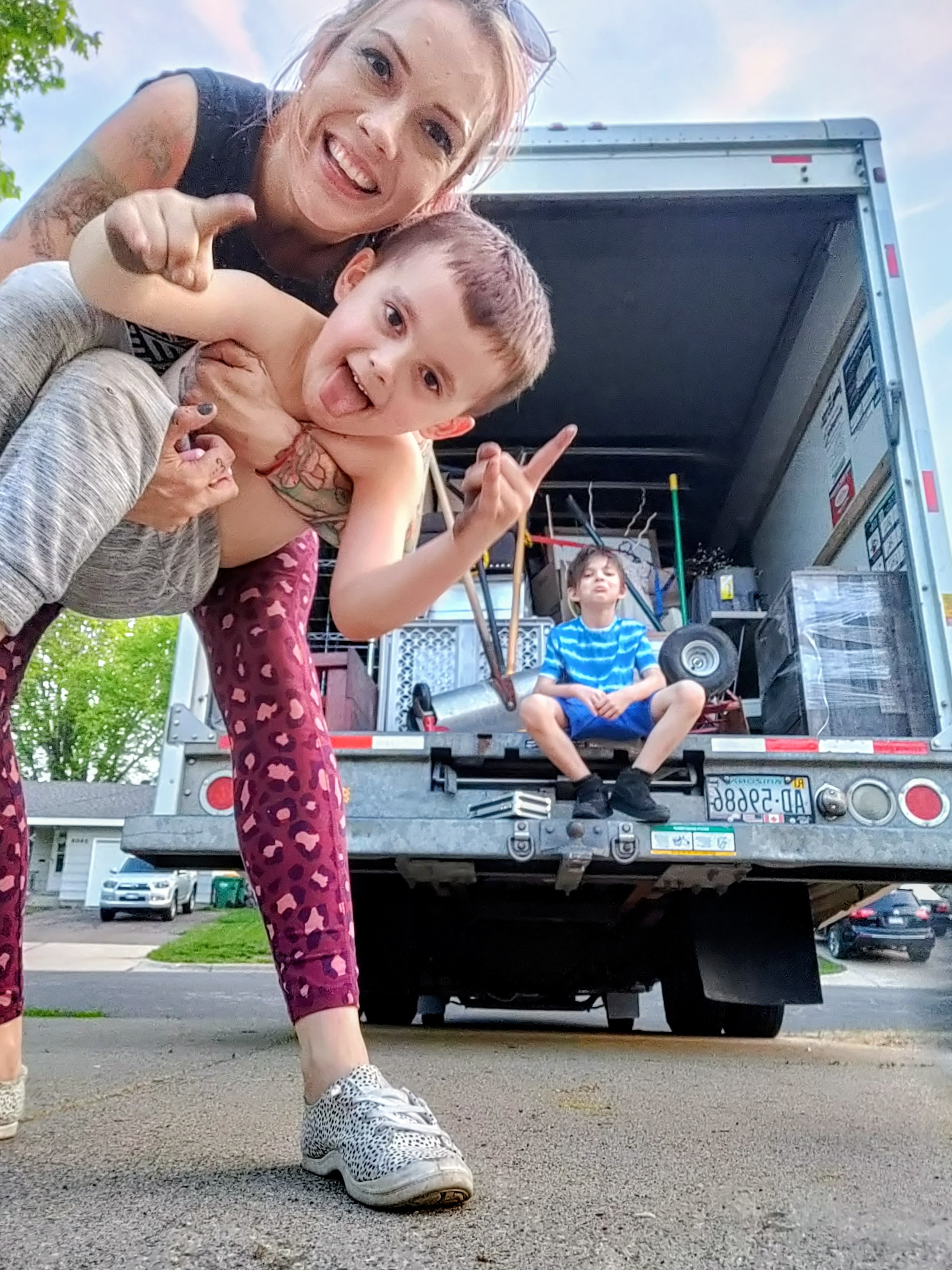 Smiling woman holds a silly boy in front of a moving truck filled with boxes.